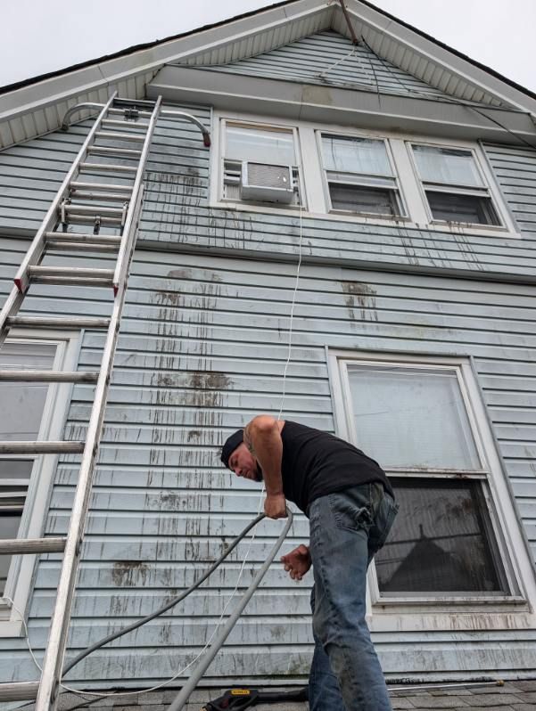 Worker on a ladder painting the side of a gray house near a window.