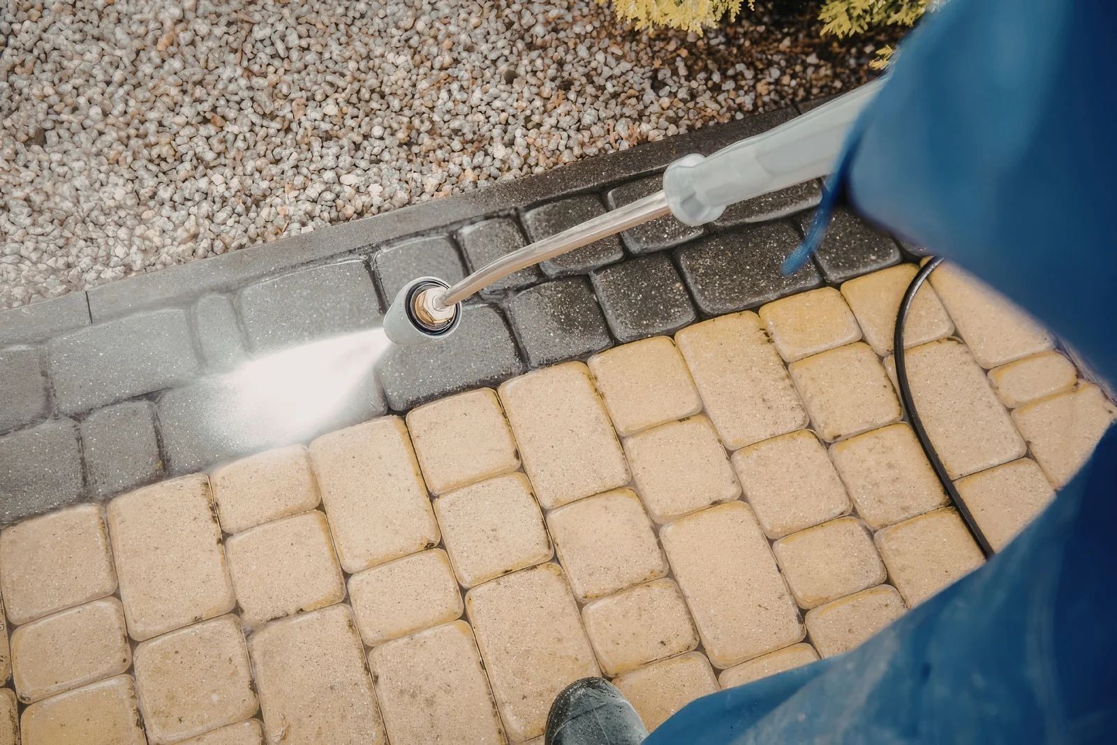 Person pressure-washing a gray walkway beside tan pavers and gravel.