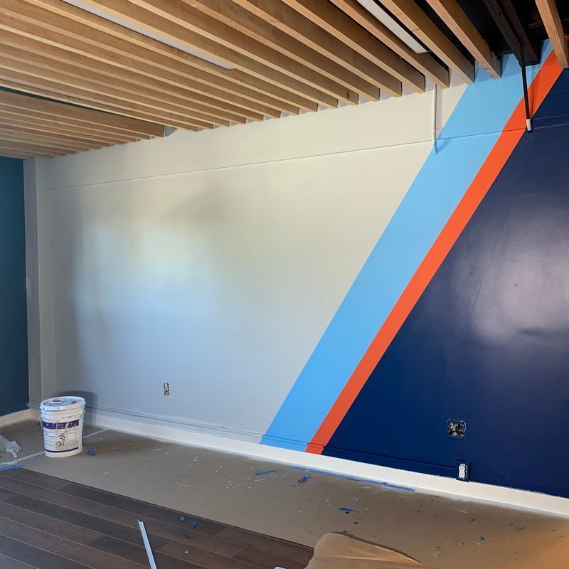 Partially painted room with blue geometric wall mural, wood slat ceiling, and paint bucket on the floor