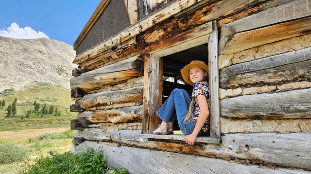 Child sitting in a cabin window, wearing a straw hat, with mountains and meadow in the background