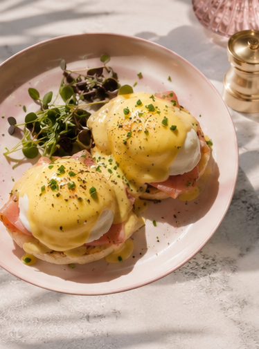 Twee Eggs Benedict op een roze bord met groene blaadjes en zwarte peper, op een zonnige tafel.