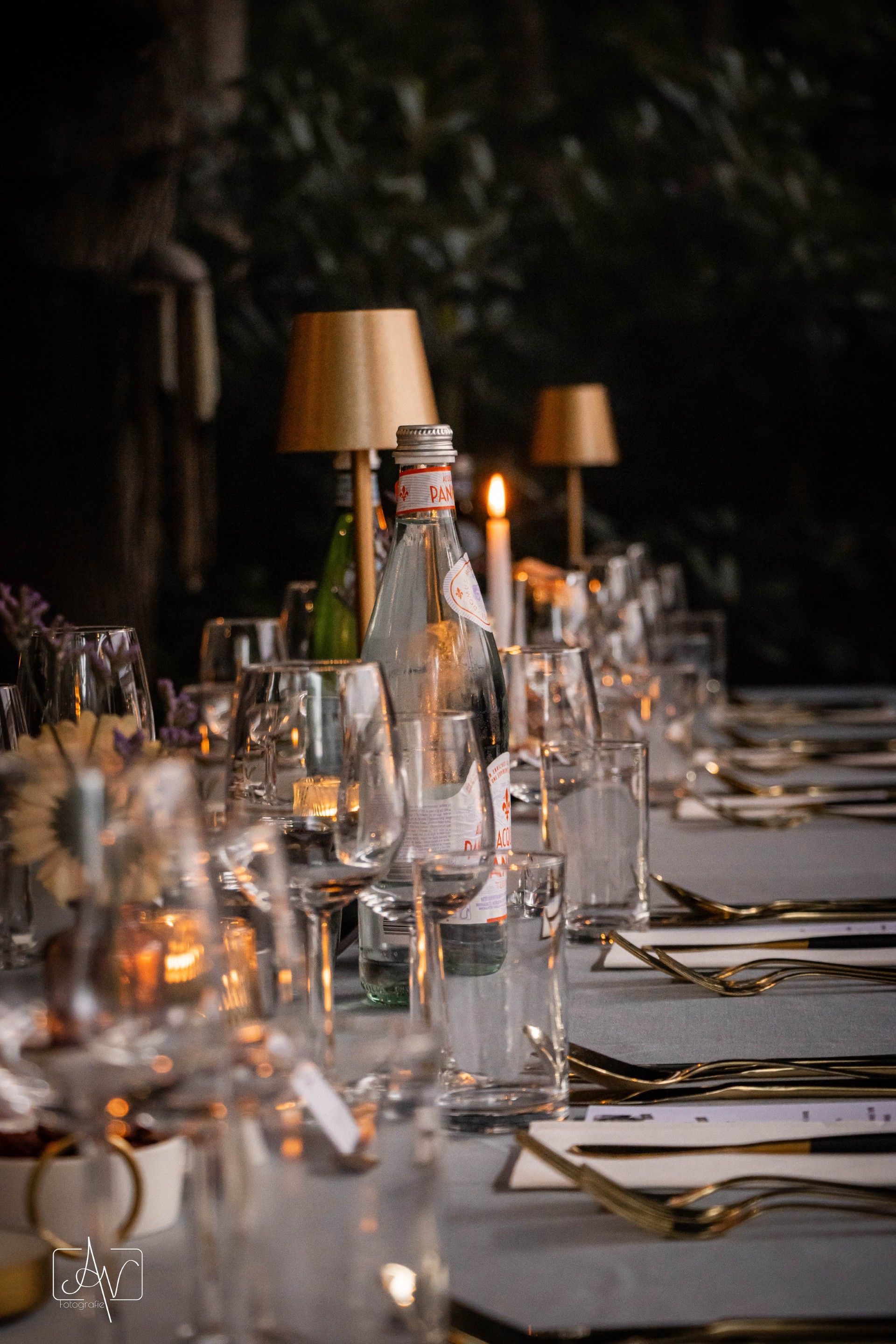 Een elegant gedekte eettafel bij kaarslicht, met glazen, gouden bestek en sfeervolle tafellampen.
