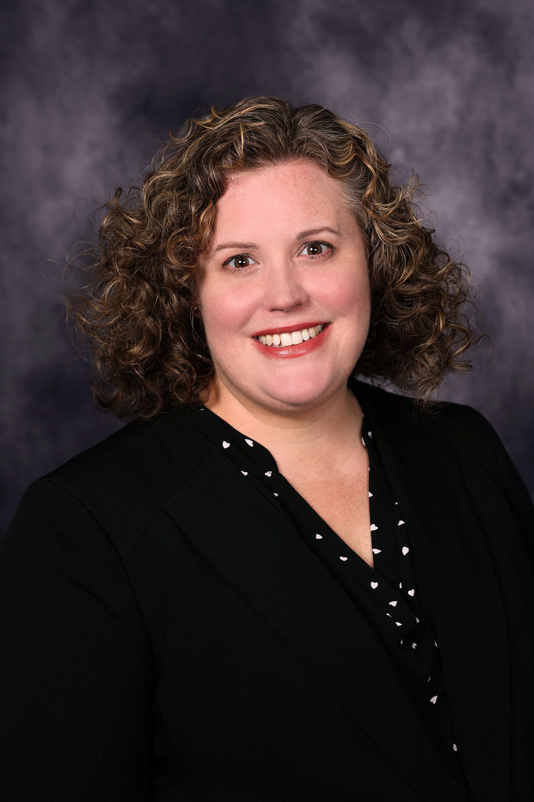 Woman with curly light brown hair smiles, wearing a gray blazer over a lavender top.