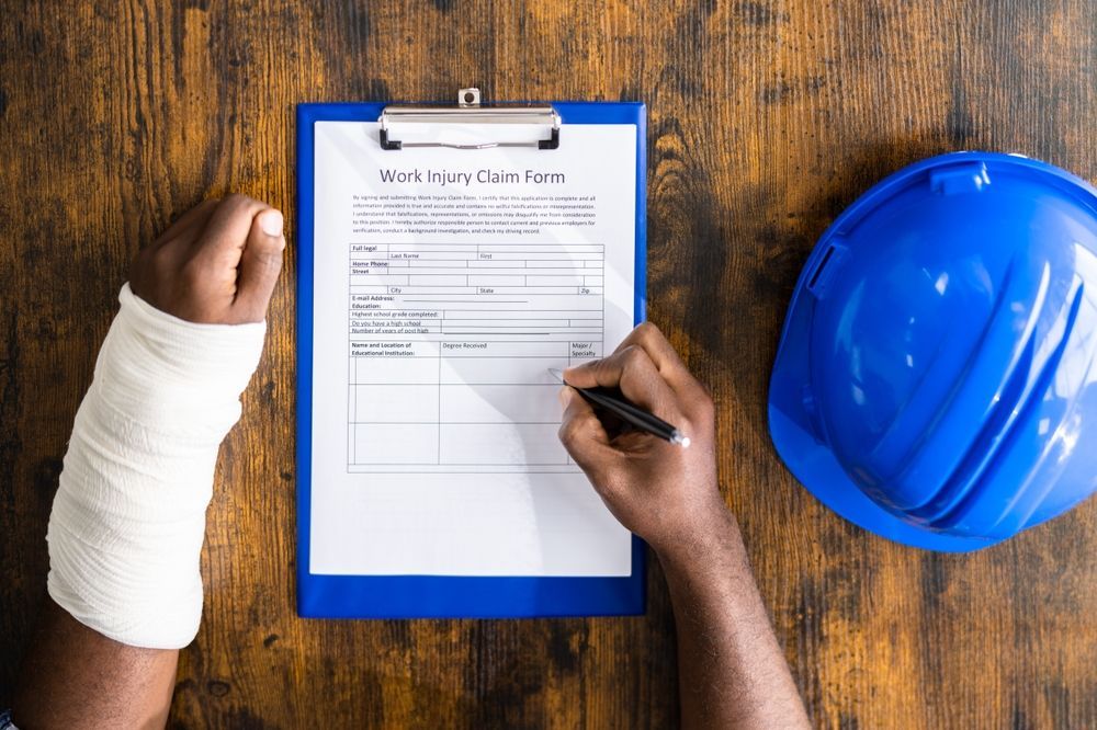Person with a wrist cast filling out a work injury claim form, blue hard hat nearby.
