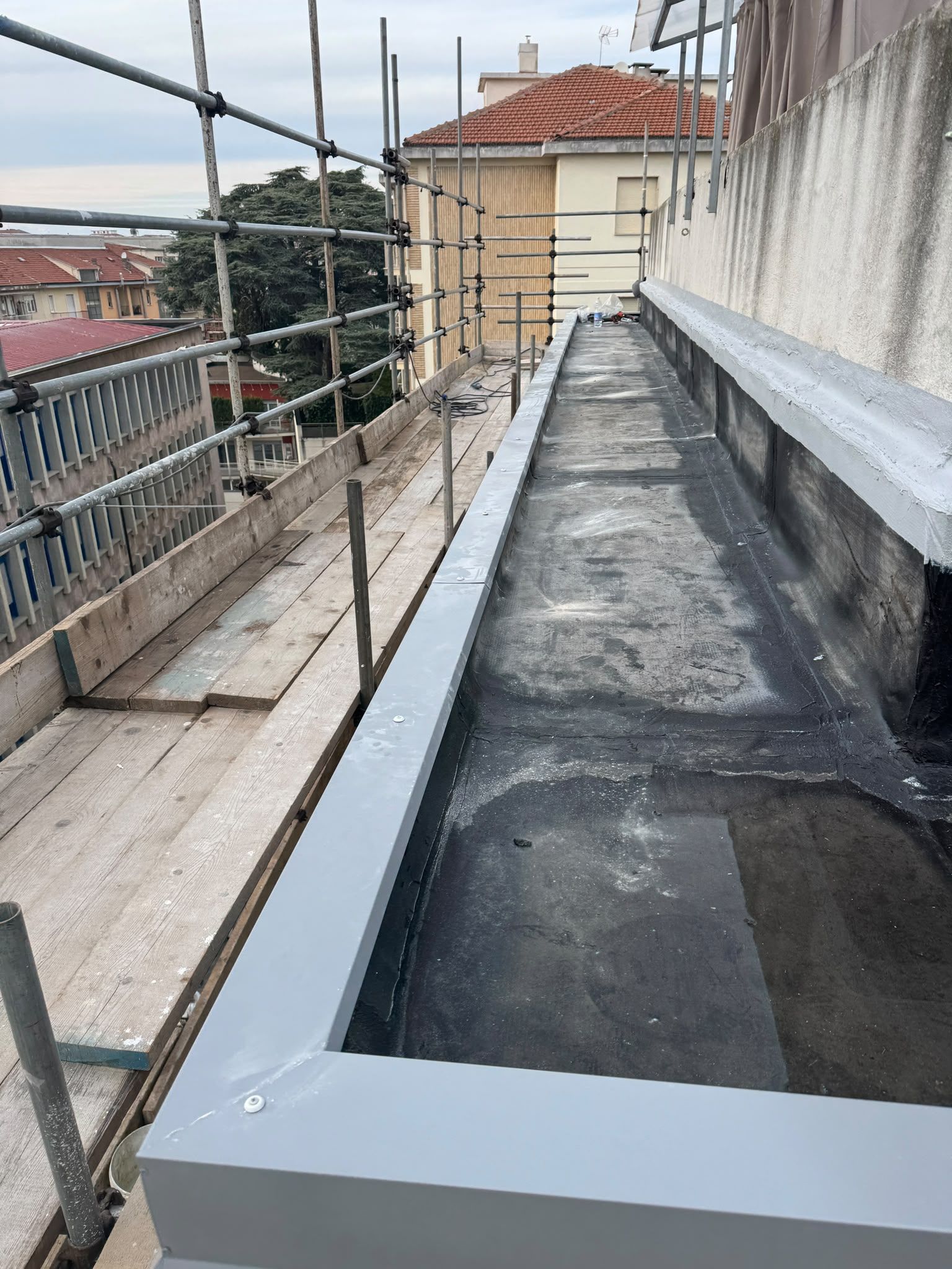 Passerella sul tetto con impalcature accanto a un edificio in costruzione, con il bordo piatto del tetto nero in evidenza.