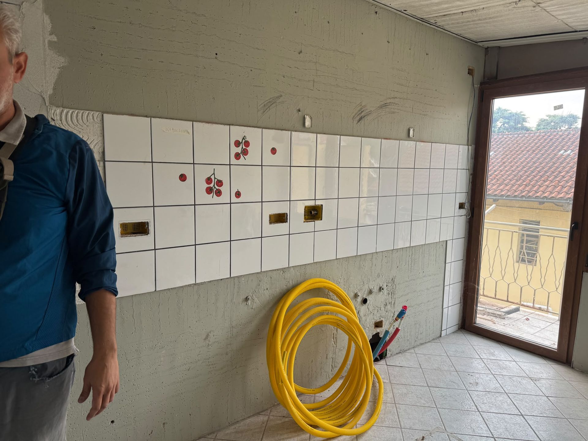 Parete parzialmente piastrellata in una stanza non finita, con un tubo giallo arrotolato e una porta che dà sull’esterno.
