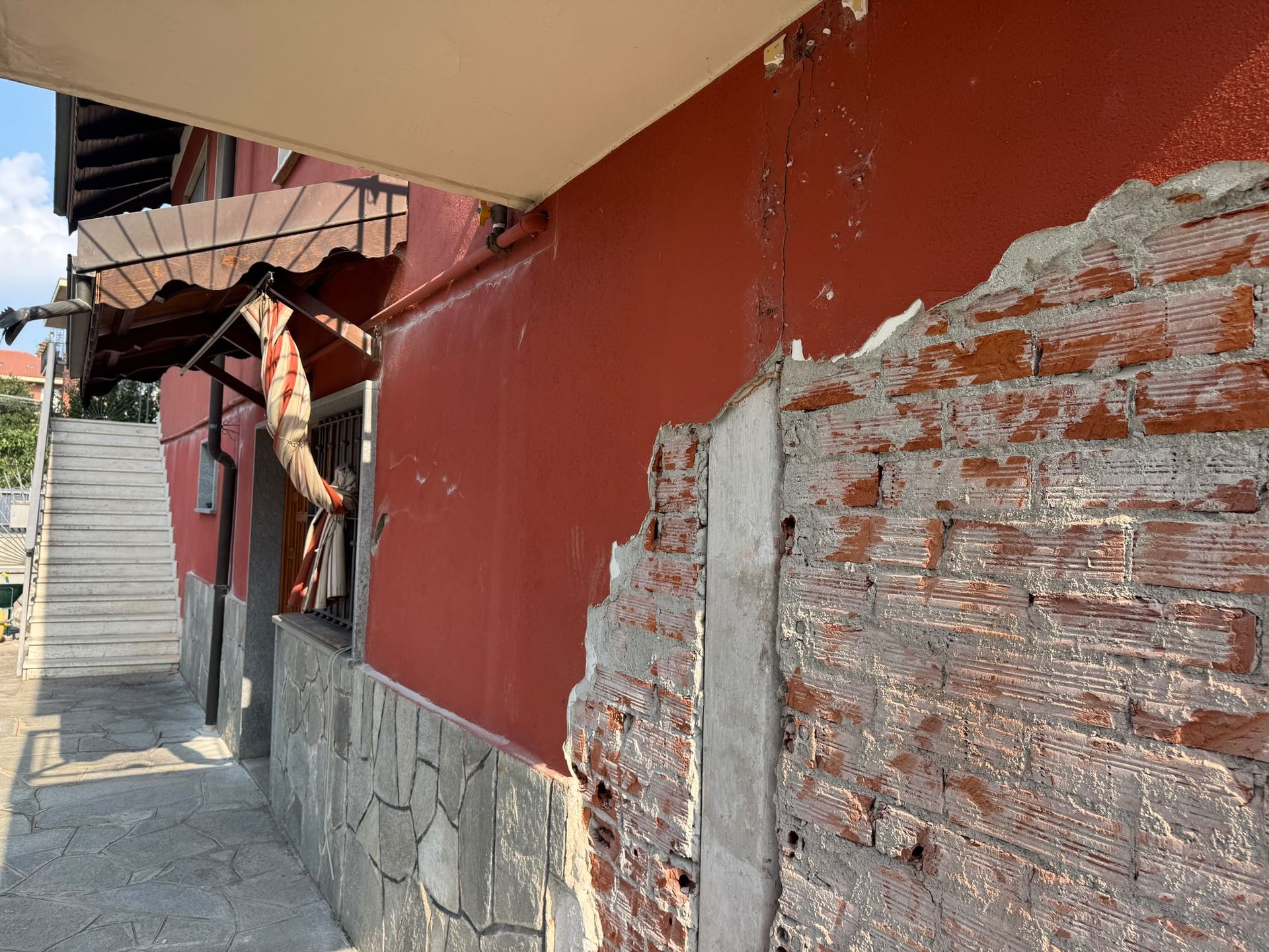 Muro rosso danneggiato, con intonaco scrostato e mattoni a vista, accanto a una scala esterna