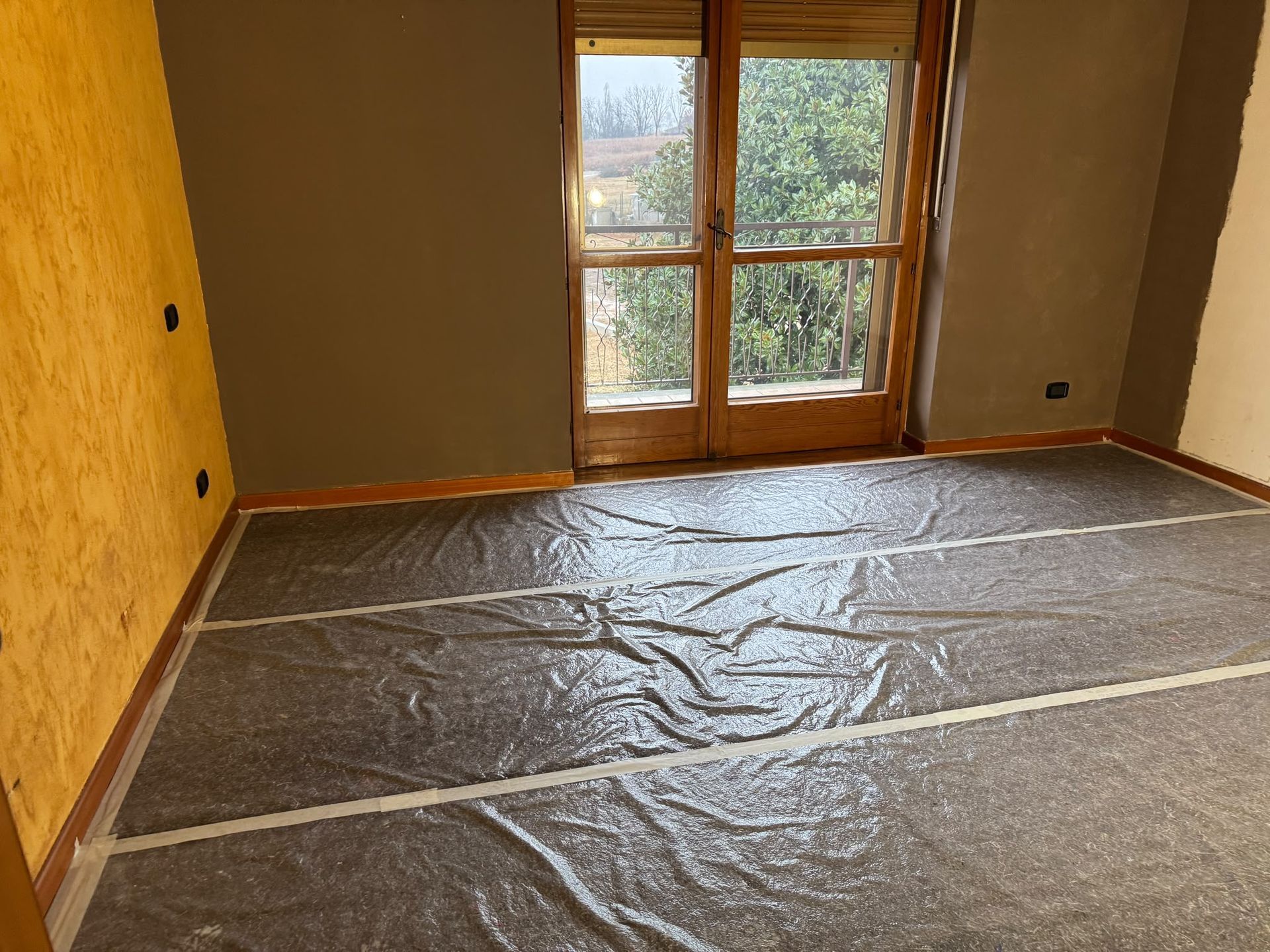 Stanza vuota con il pavimento coperto di plastica, pareti color beige e una finestra da cui entra la luce del giorno