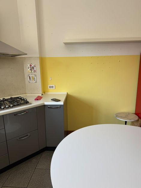Piccola cucina con pareti gialle, mobili grigi, piano cottura, lavello e un tavolo rotondo bianco in primo piano