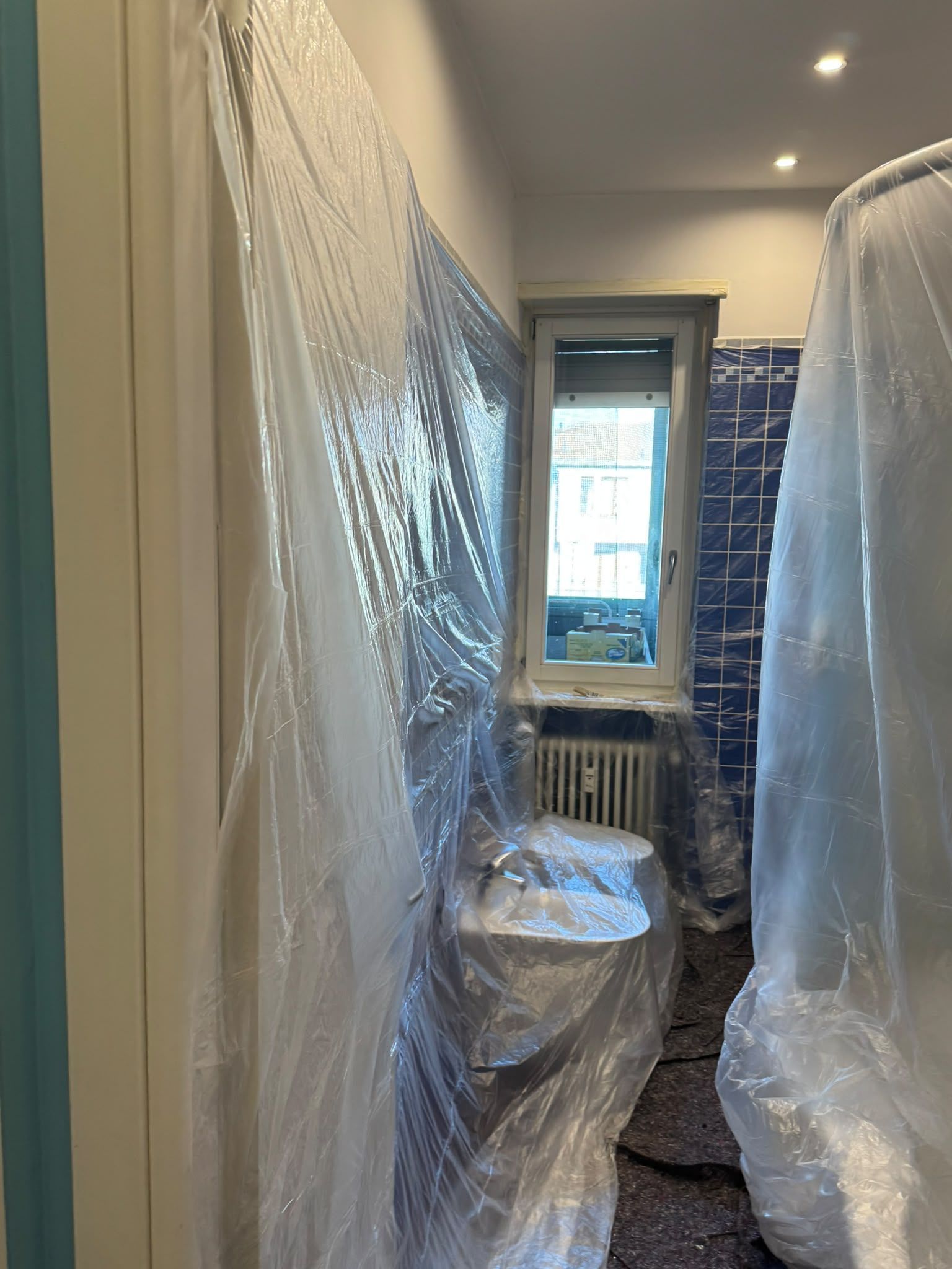 Bagno stretto in fase di ristrutturazione, con i sanitari avvolti nella plastica e una finestra in fondo.
