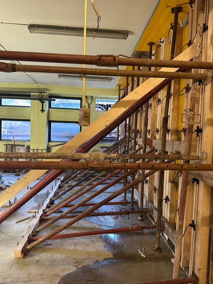 Interno di un cantiere edile con impalcature in legno, puntelli e struttura a vista lungo un pavimento bagnato.