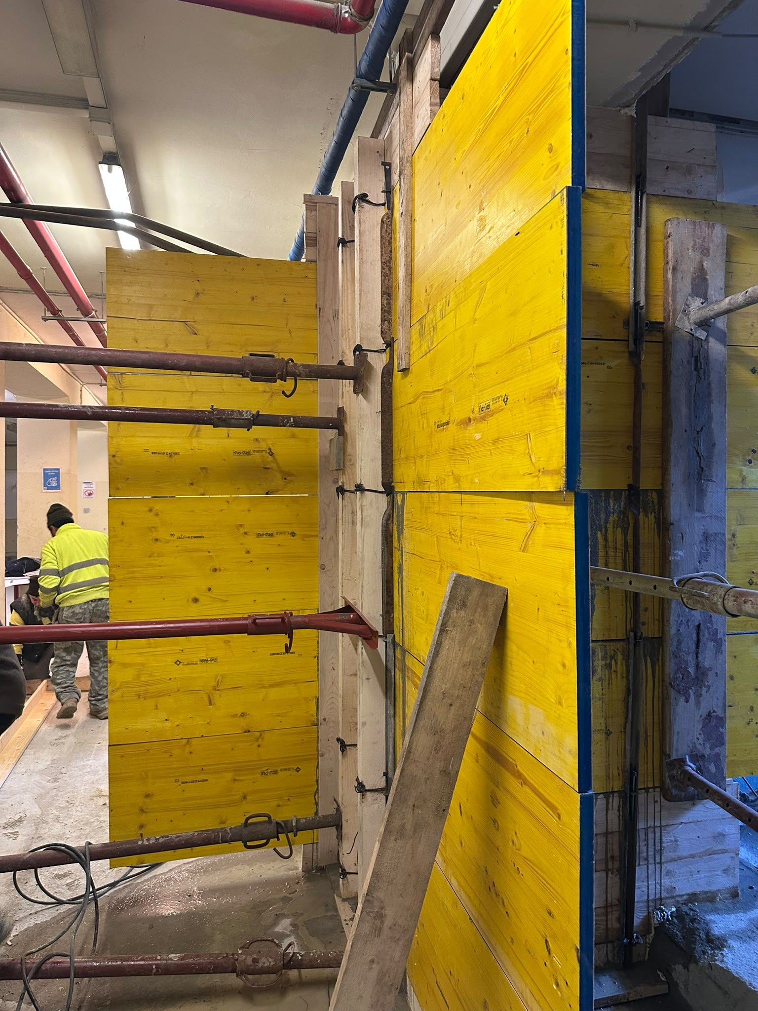 Pannelli gialli per casseforme in un cantiere, con puntelli d’acciaio e un operaio con casco protettivo sullo sfondo