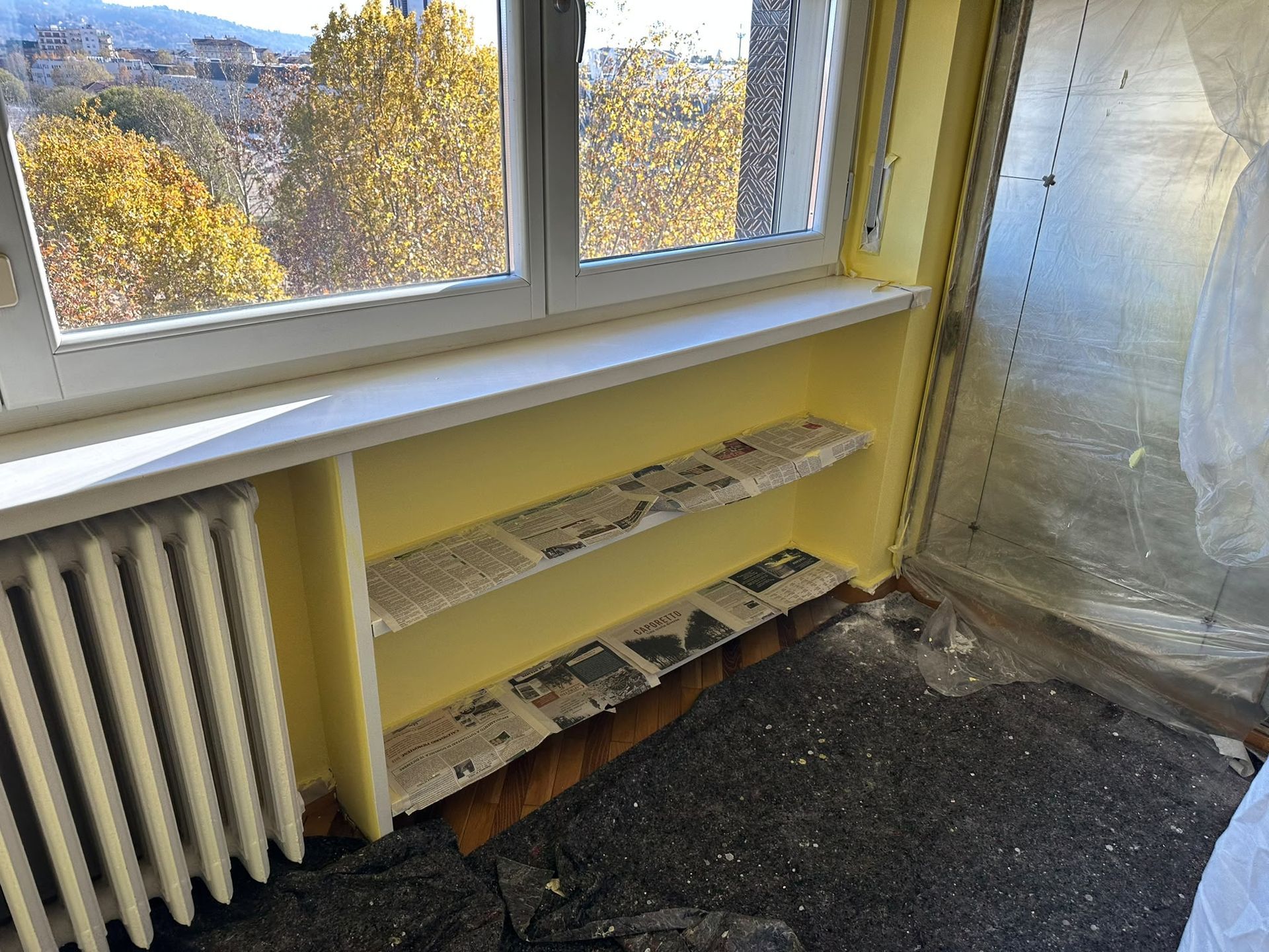 Angolo di una stanza vuota con una finestra, un termosifone, scaffali gialli e una parete coperta di plastica