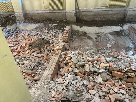 Pavimento parzialmente demolito, con macerie, mattoni e detriti di cemento accanto a un muro e a una recinzione