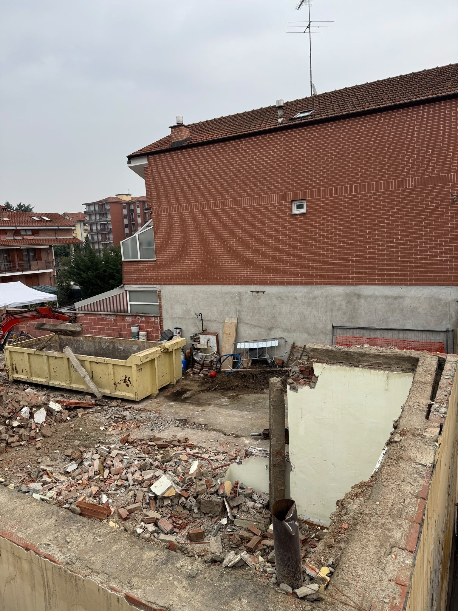 Cantiere di demolizione cosparso di macerie accanto a un edificio con il tetto di tegole rosse, sotto un cielo coperto