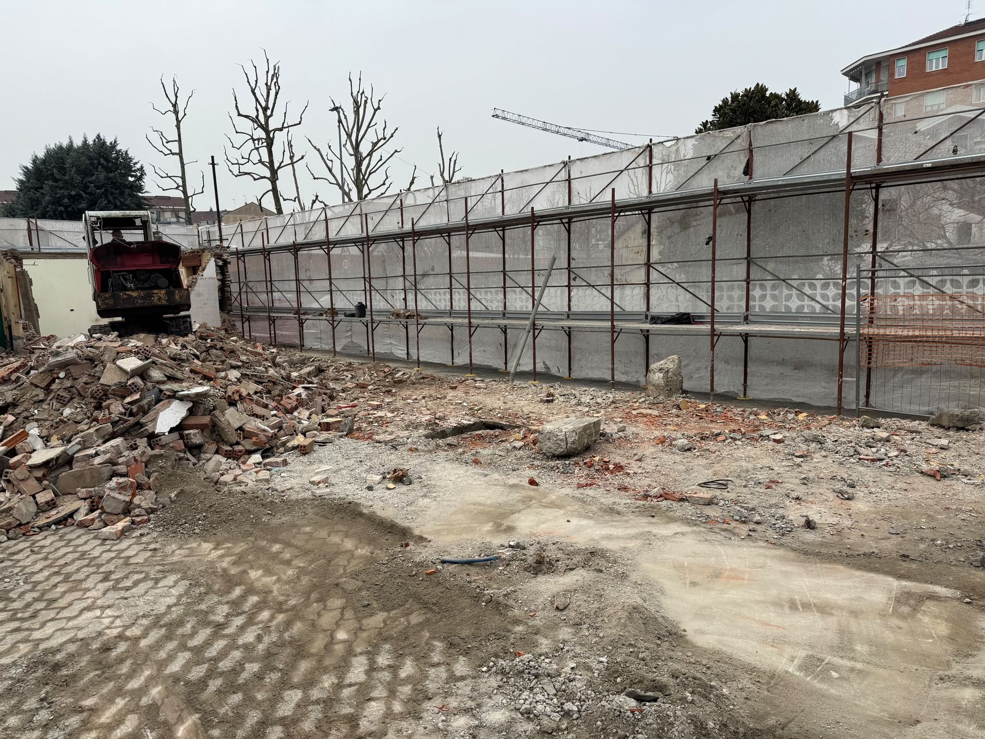 Cantiere edile con macerie, un camion rosso e un lungo muro di cemento sotto un’impalcatura