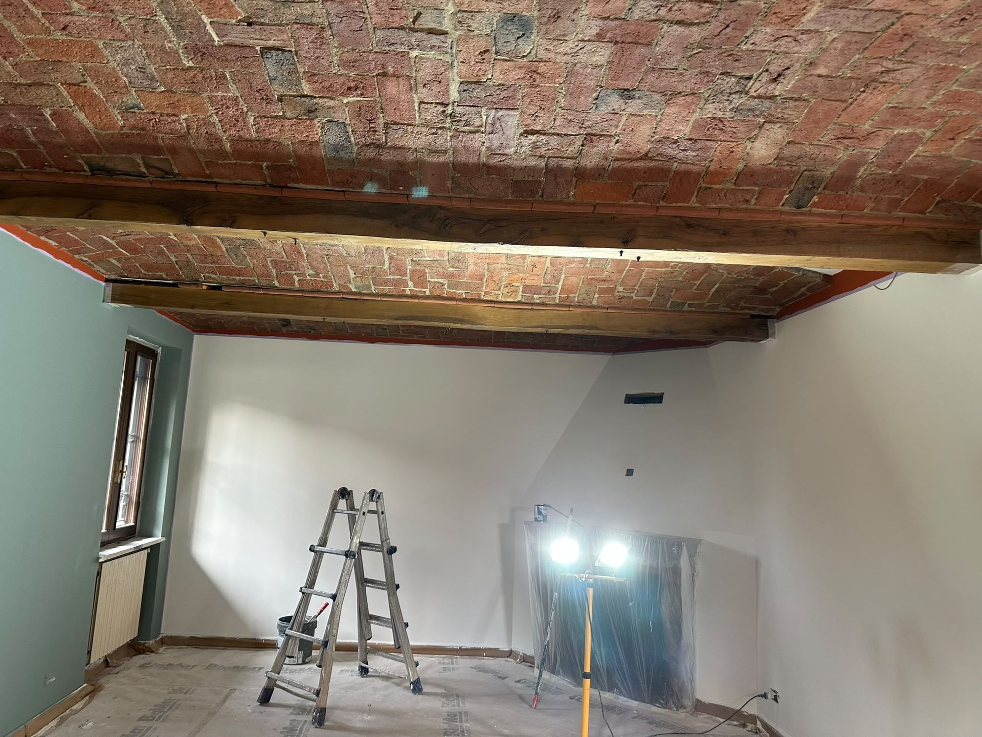Stanza in ristrutturazione con soffitto in mattoni a vista, scala, specchio e luci da lavoro intense vicino a una finestra