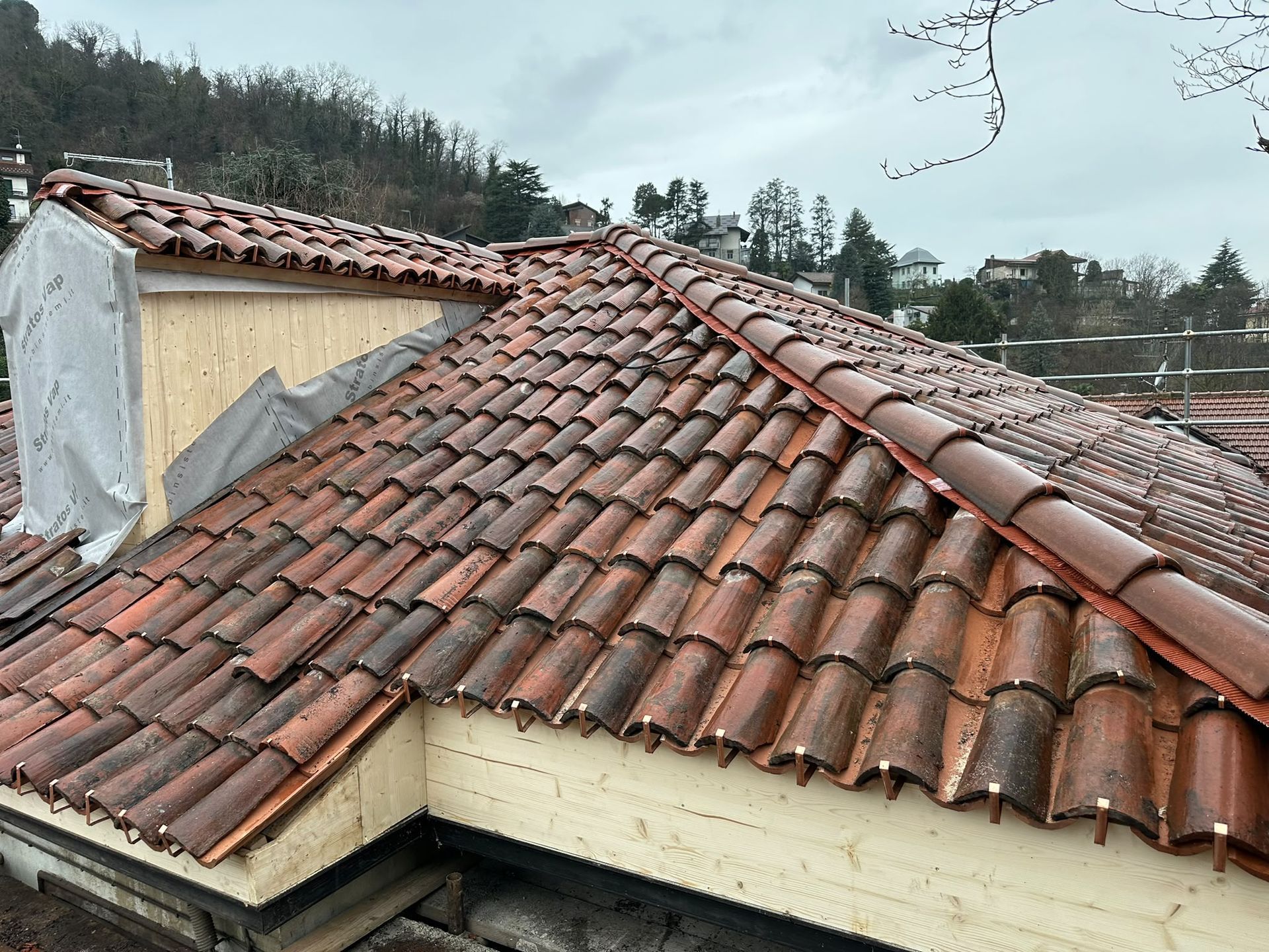 Tetto in tegole di argilla rossa con un bordo danneggiato e una sezione parzialmente coperta, sotto un cielo coperto
