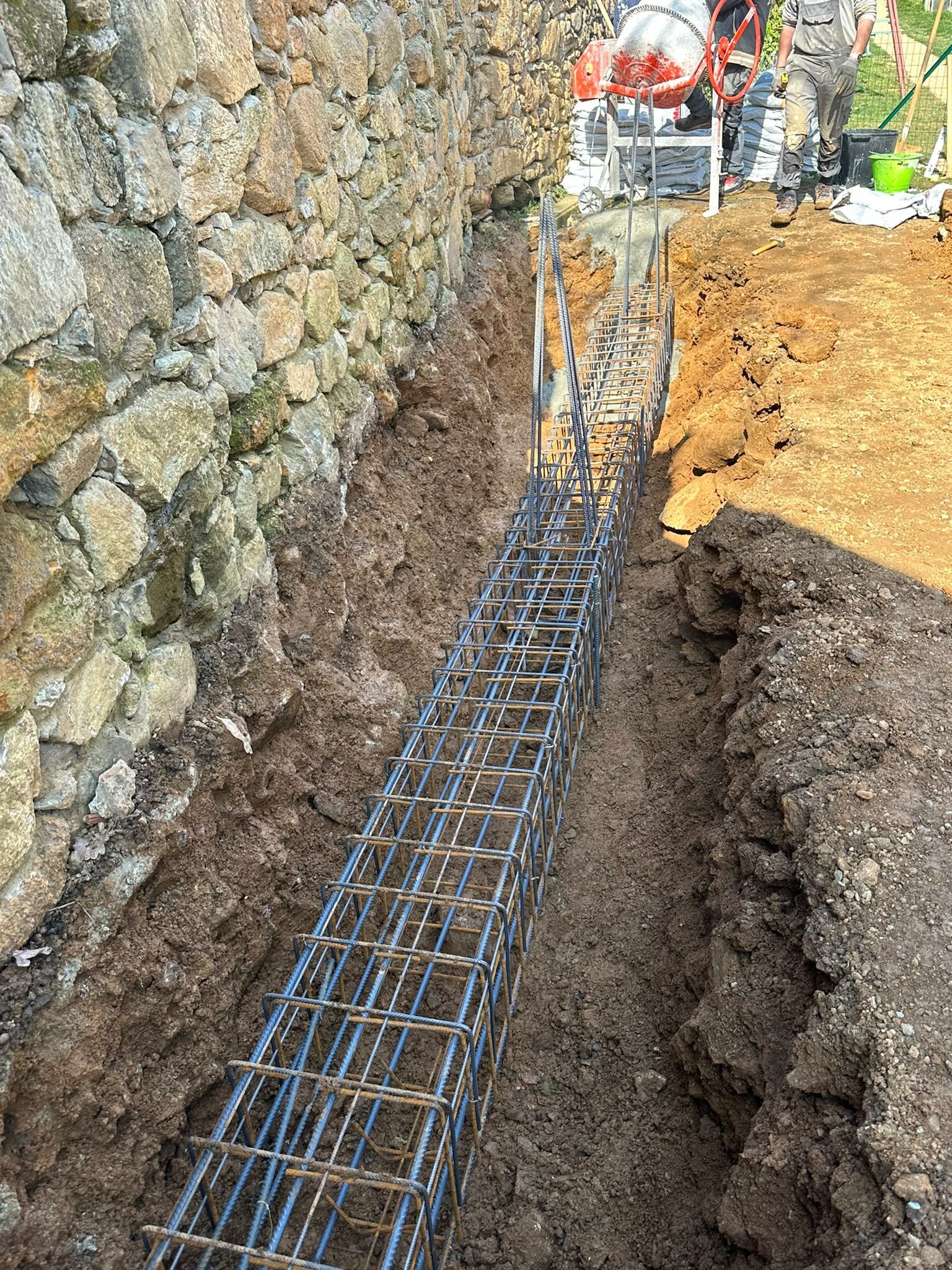 Trincea con gabbia d’armatura in acciaio accanto a un muro di pietra in un cantiere edile