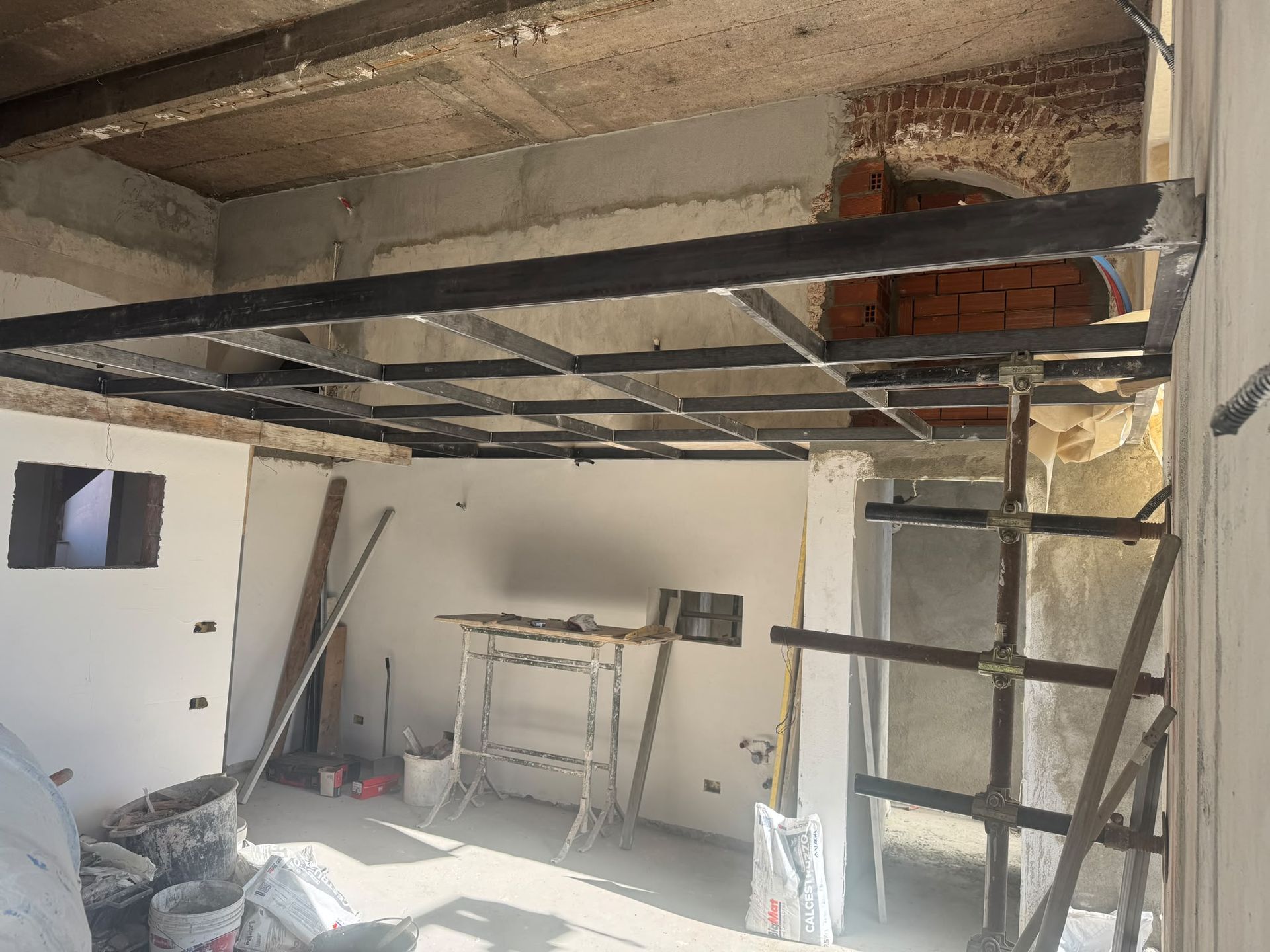 Cantiere di ristrutturazione con impalcature metalliche sotto un soffitto parzialmente scoperto e una parete in mattoni.