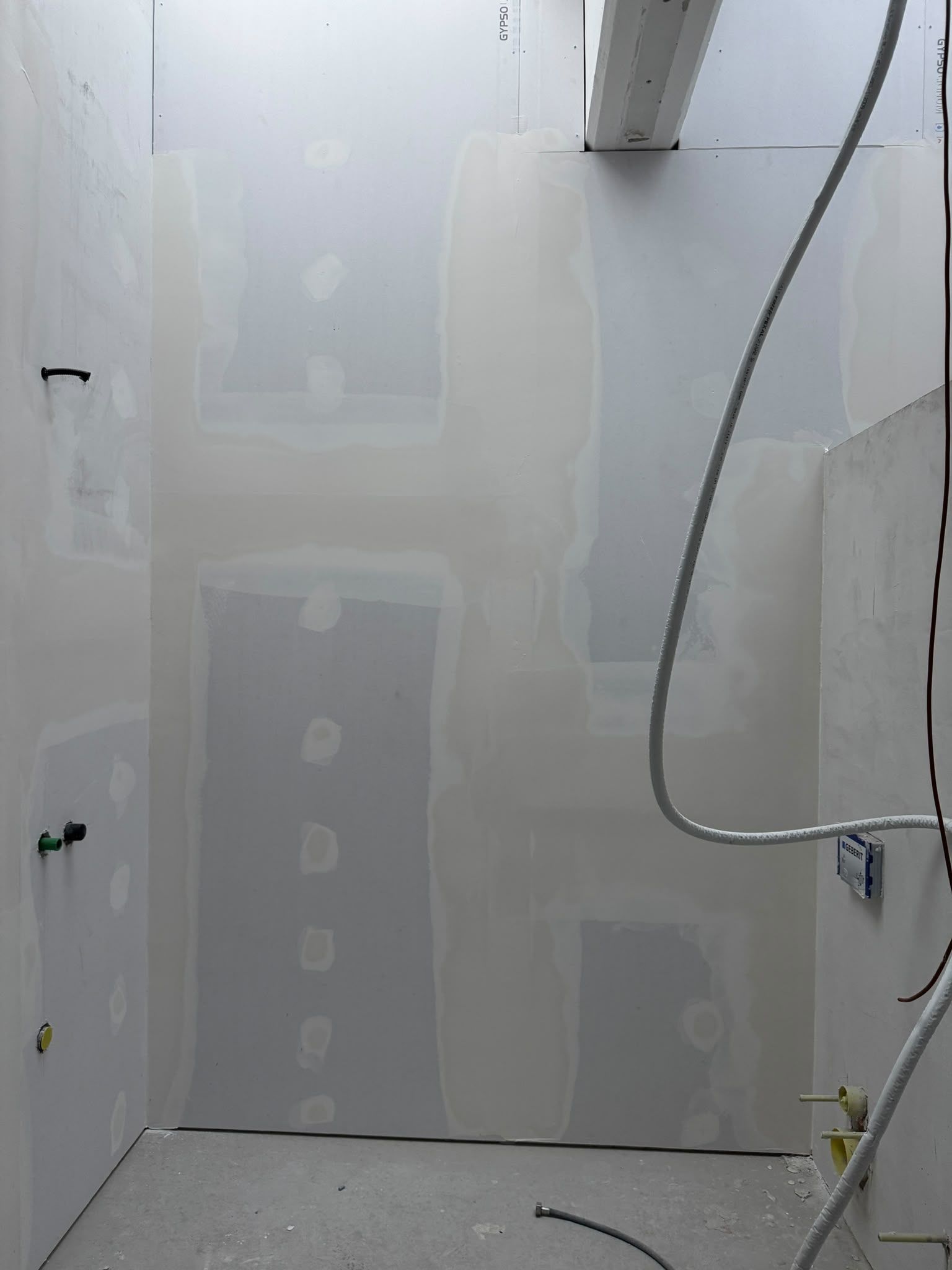 Bagno incompleto con rappezzi di cartongesso, tubi a vista e un flessibile della doccia penzolante.