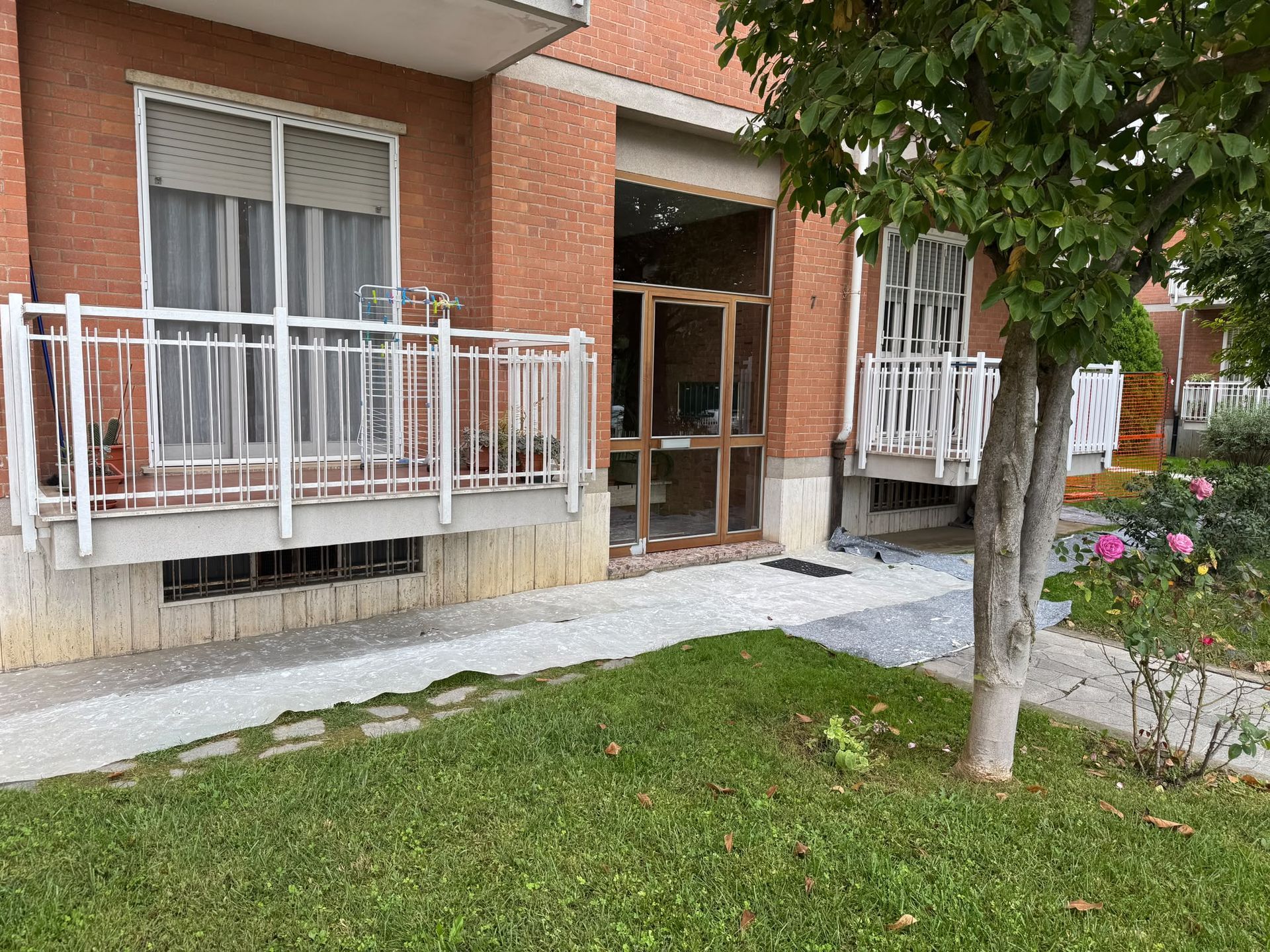 Ingresso di un condominio con ringhiere bianche, porta a vetri e un piccolo prato con un albero davanti