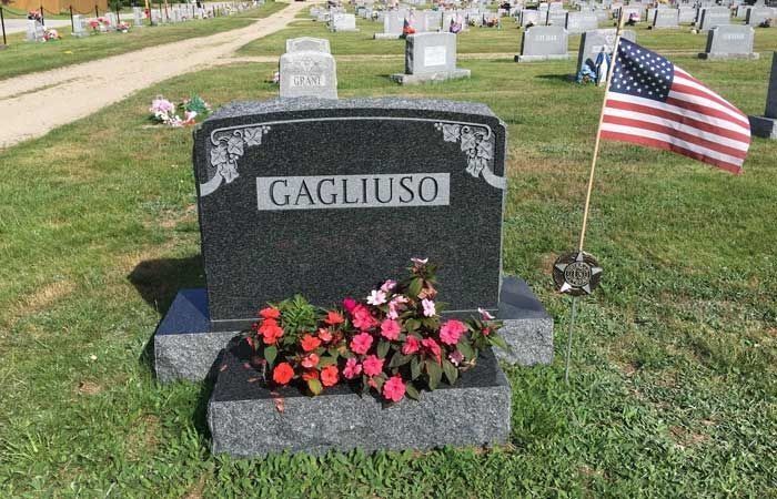 Gravestone marked “CAGLIUSO” with red flowers and an American flag in a cemetery