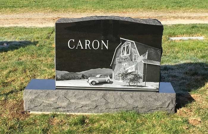 Black granite gravestone with a car and house engraving, labeled “CARON,” on a grassy lawn.
