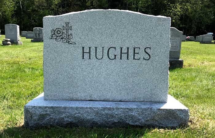 Gray granite gravestone engraved “HUGHES” in a cemetery, with trees and other markers in the background