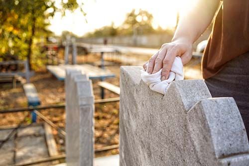 Hand wiping a gray gravestone in a sunlit cemetery