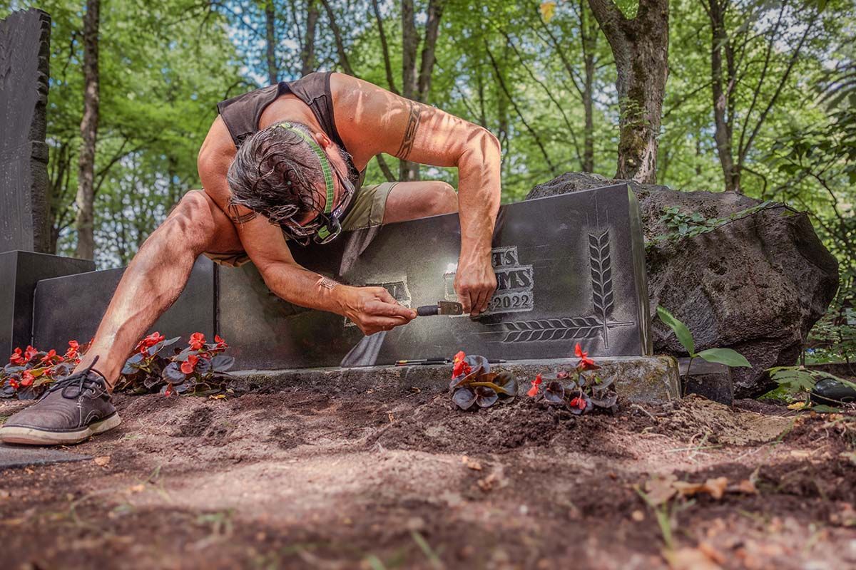 Person in muddy orange shirt kneels by a gravestone in a forested cemetery, cleaning with a brush.