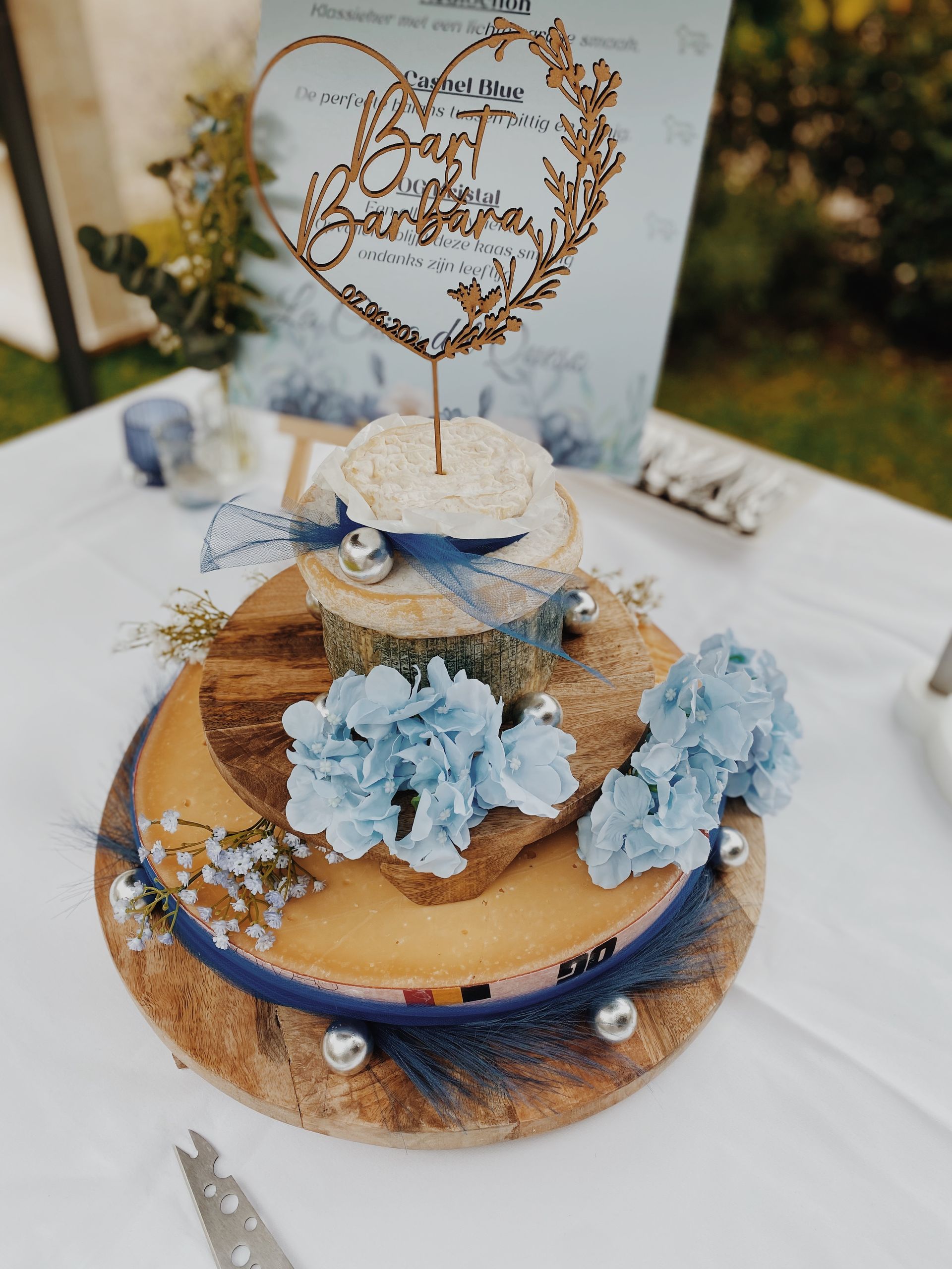 Een houten taartstandaard met twee lagen als tafeldecoratie, versierd met lichtblauwe bloemen, een donkerblauw lint en een gouden hartvormige taarttopper.