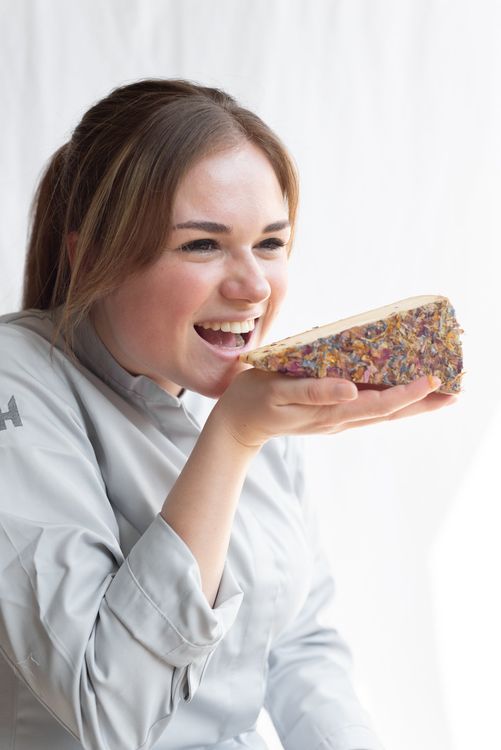 Een persoon in een grijze koksjas lacht en houdt een stuk taart vast dat bedekt is met veelkleurige hagelslag.