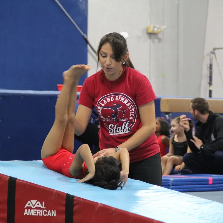 A woman in a red shirt that says land gymnastics is helping a young girl on a balance beam