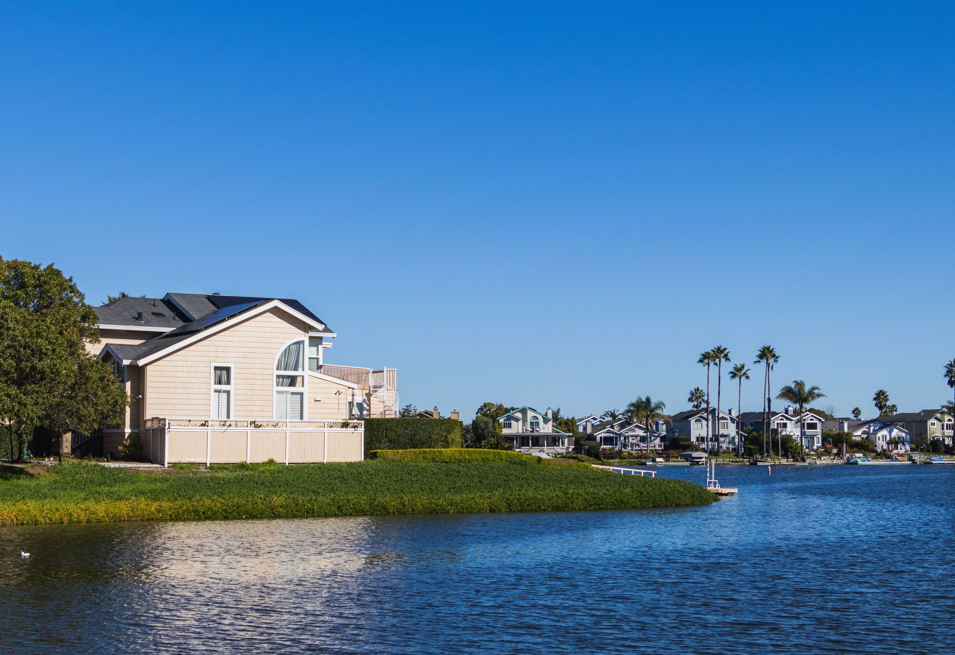 Waterfront homes beside a calm canal under a clear blue sky, with palm trees in the distance