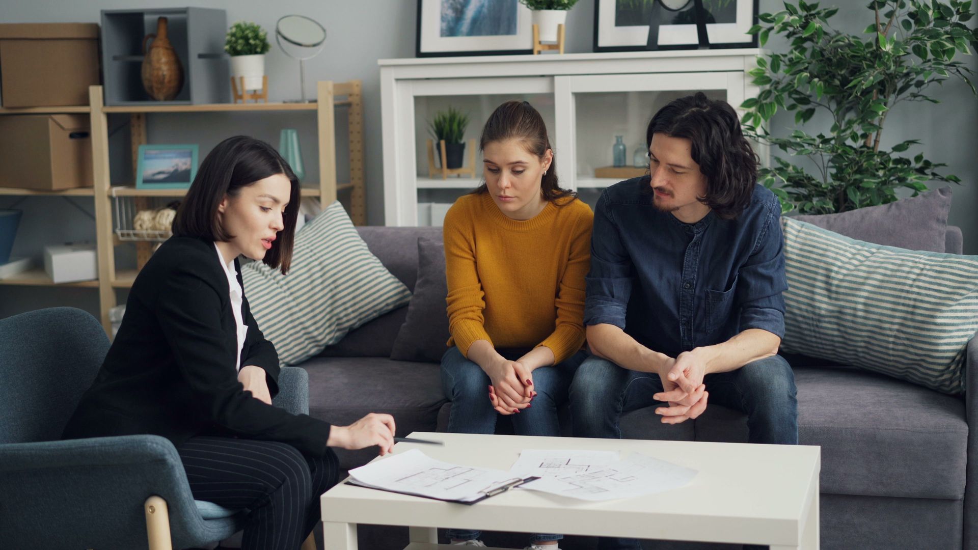 Three people sit on a couch and chair, talking with papers on a coffee table in a cozy living room.