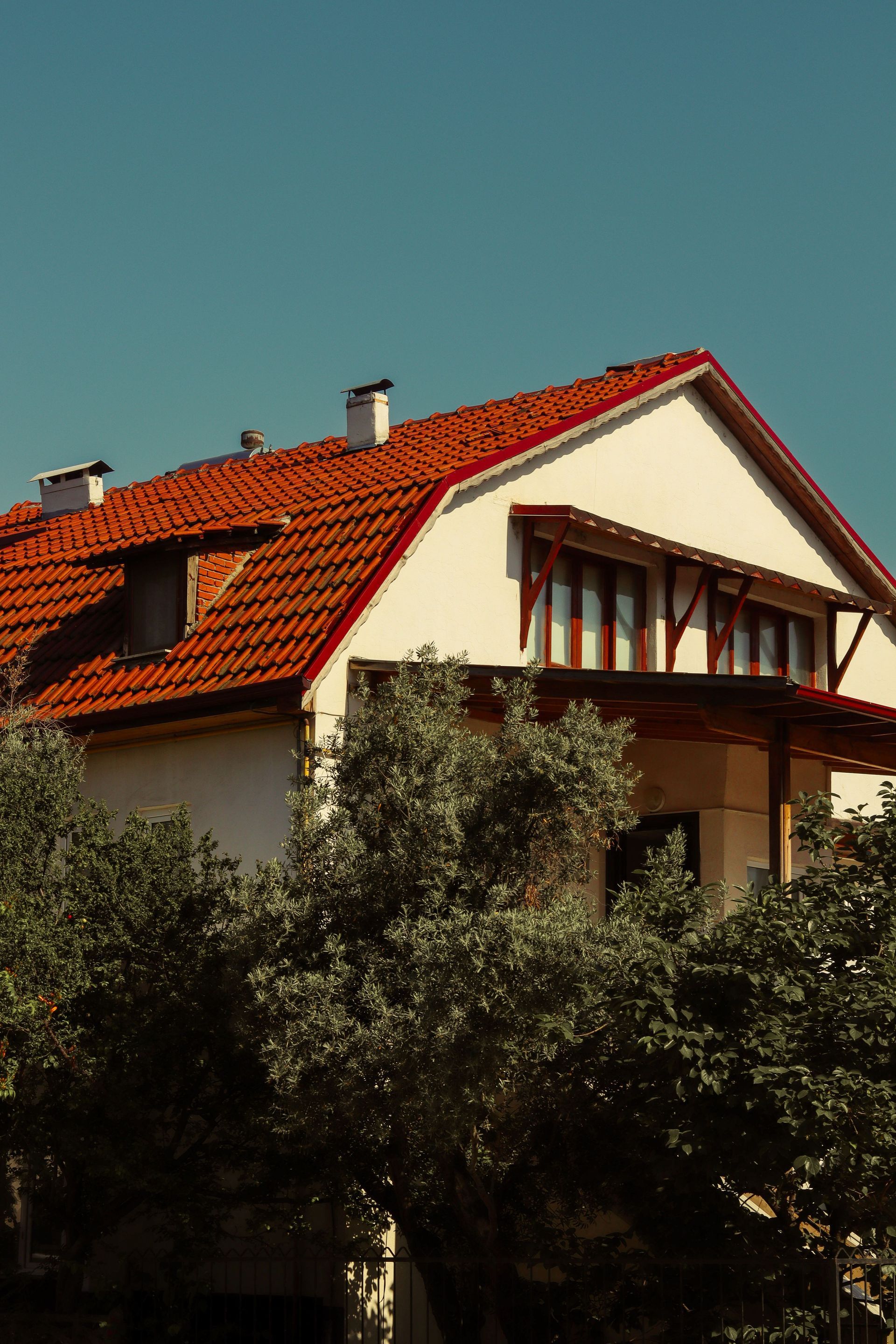 Red-roofed house with beige walls and wooden balcony behind tall trees under a clear blue sky