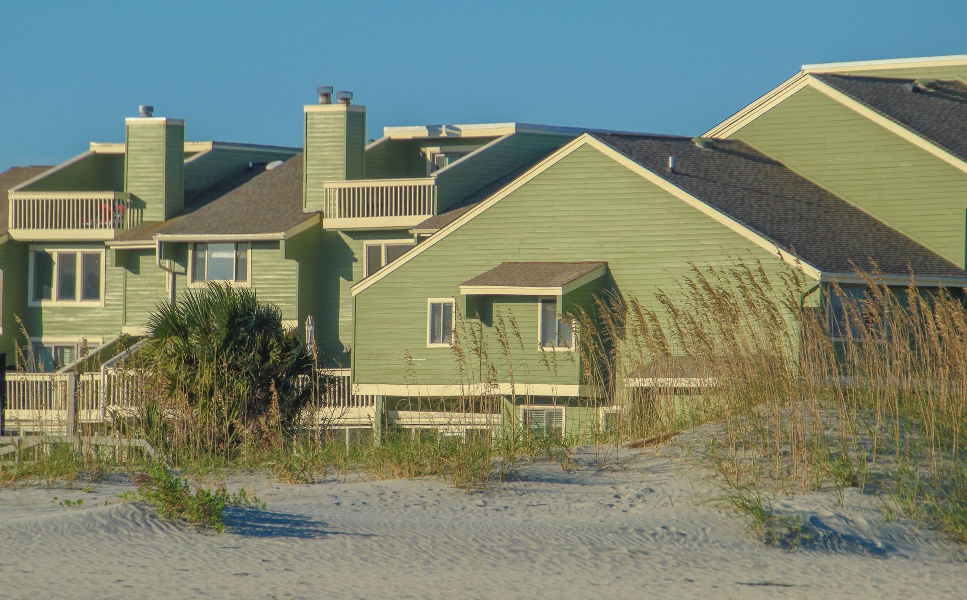 Green beachfront houses with chimneys and tall grass beside sandy dunes under clear sky