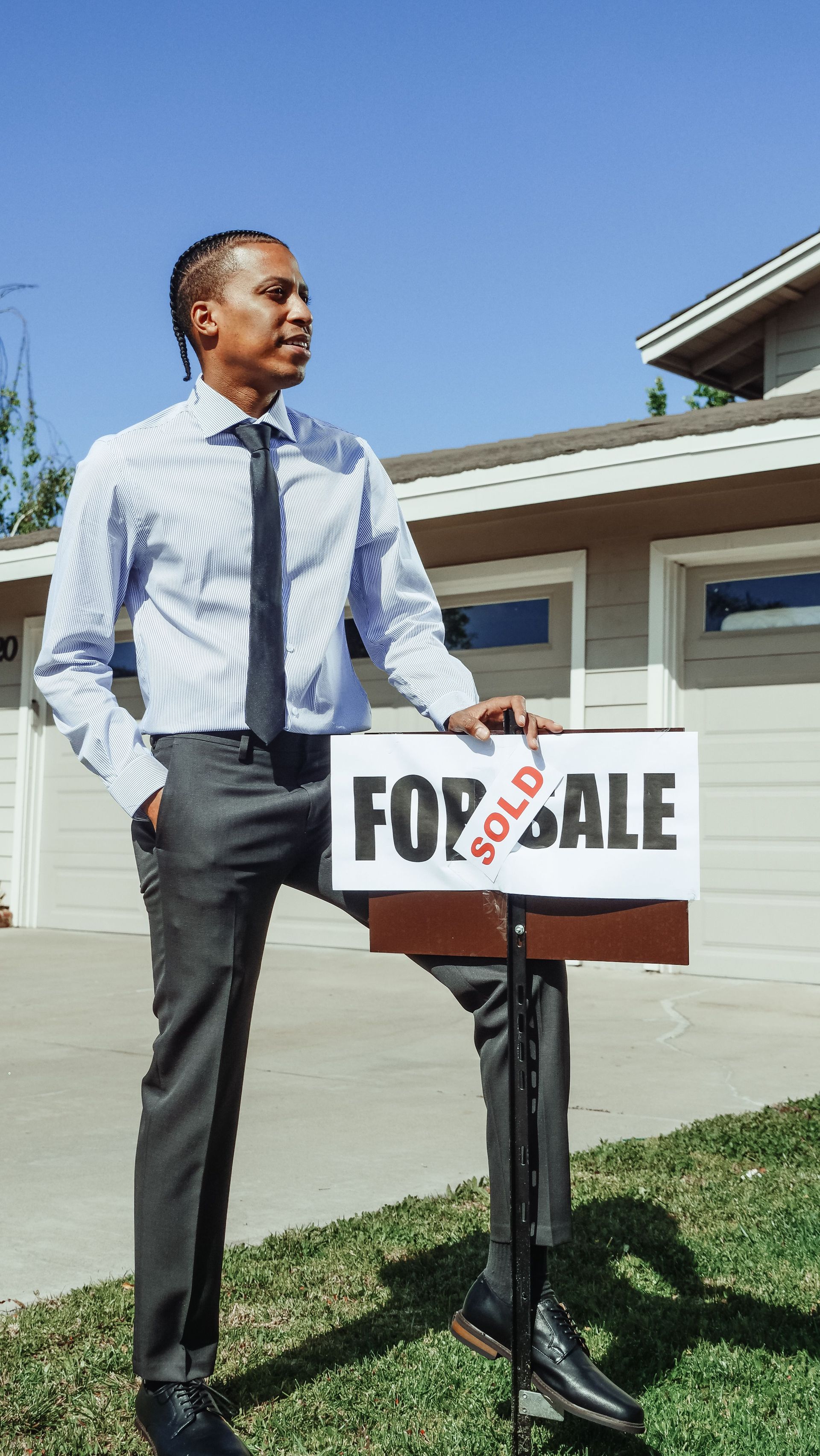 Man standing beside a house with a “For Sale” sign in the driveway under a clear blue sky