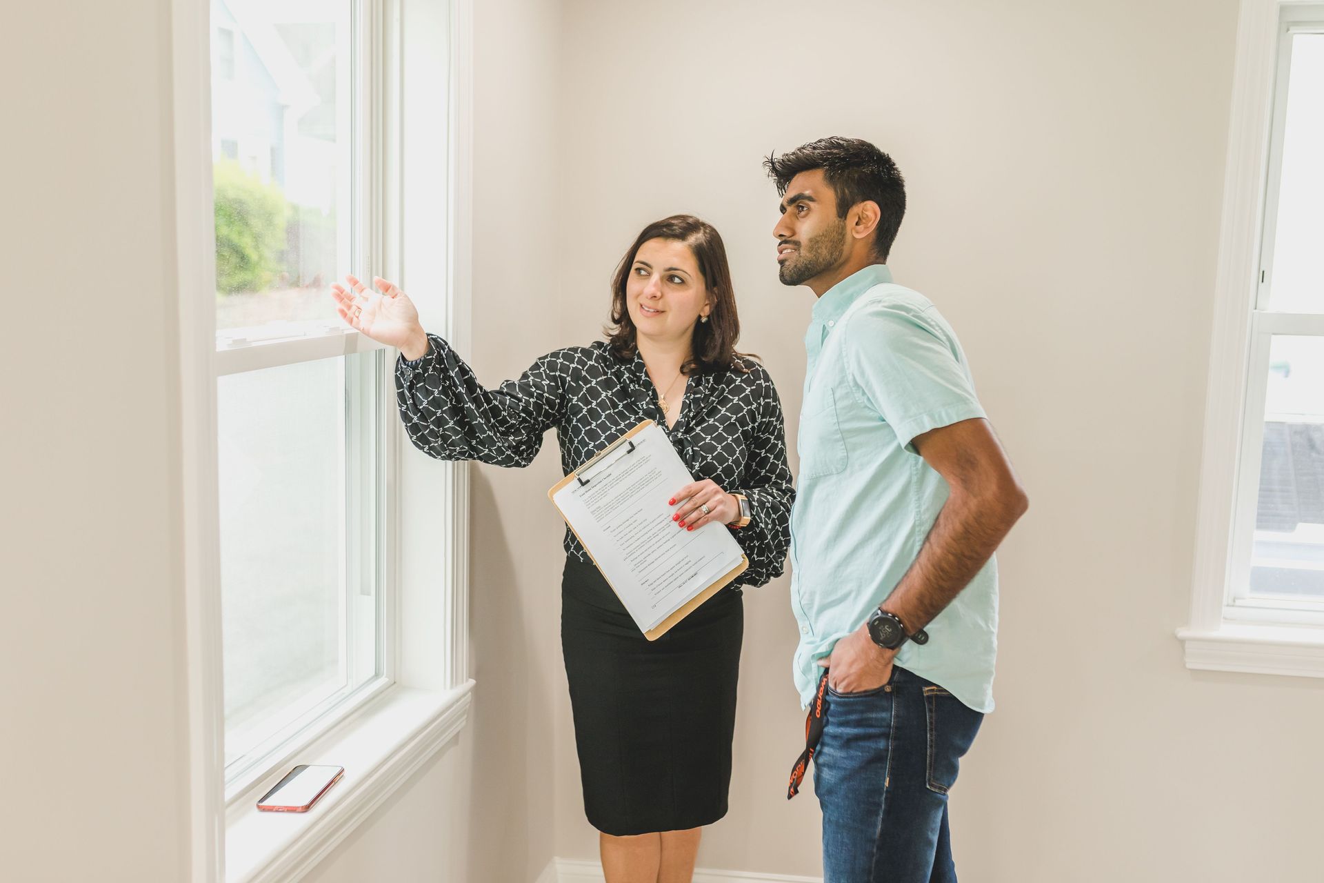 Two people discussing a room, one holding a clipboard and pointing toward a window