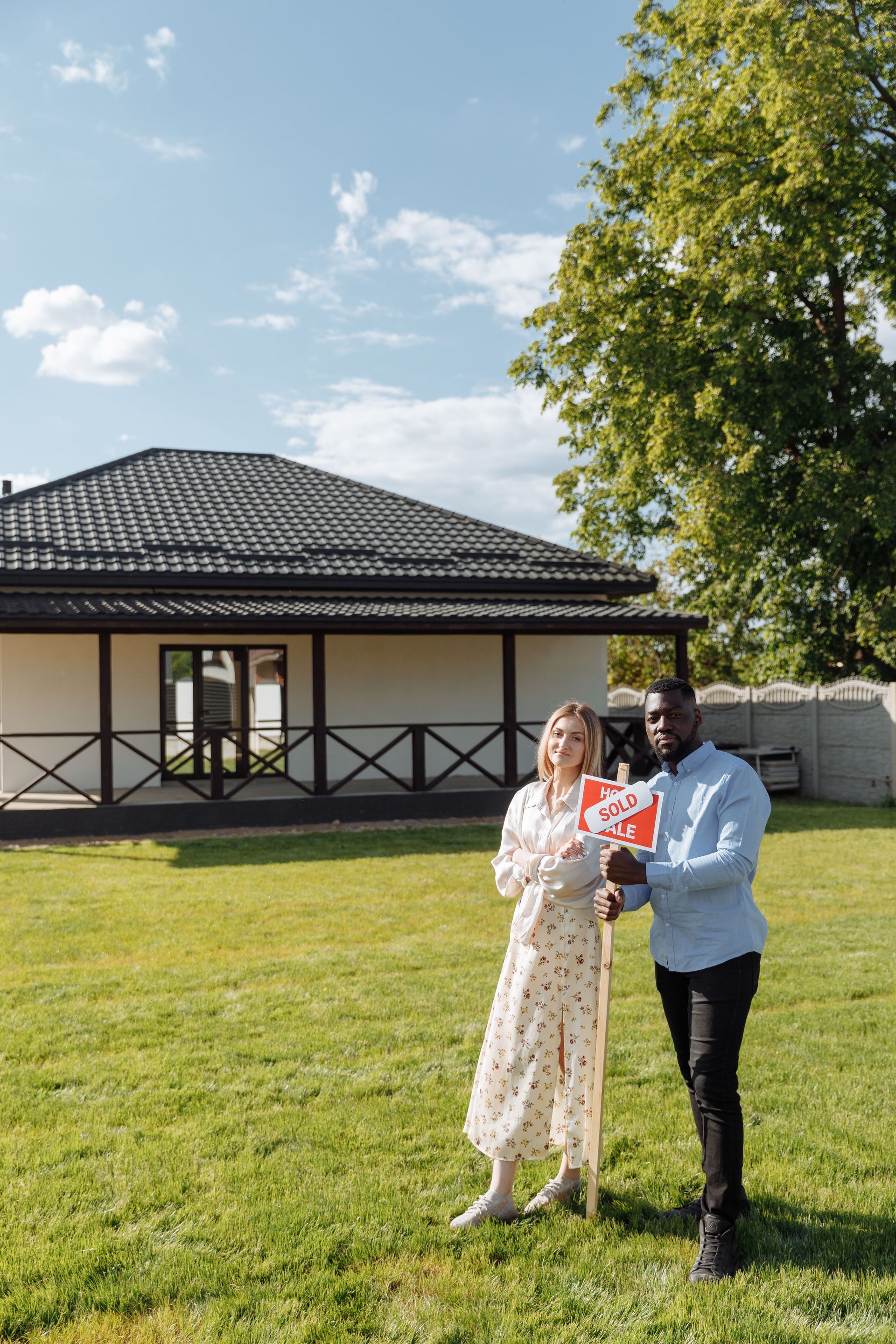 Two people standing on a lawn in front of a house, holding a red book in bright daylight