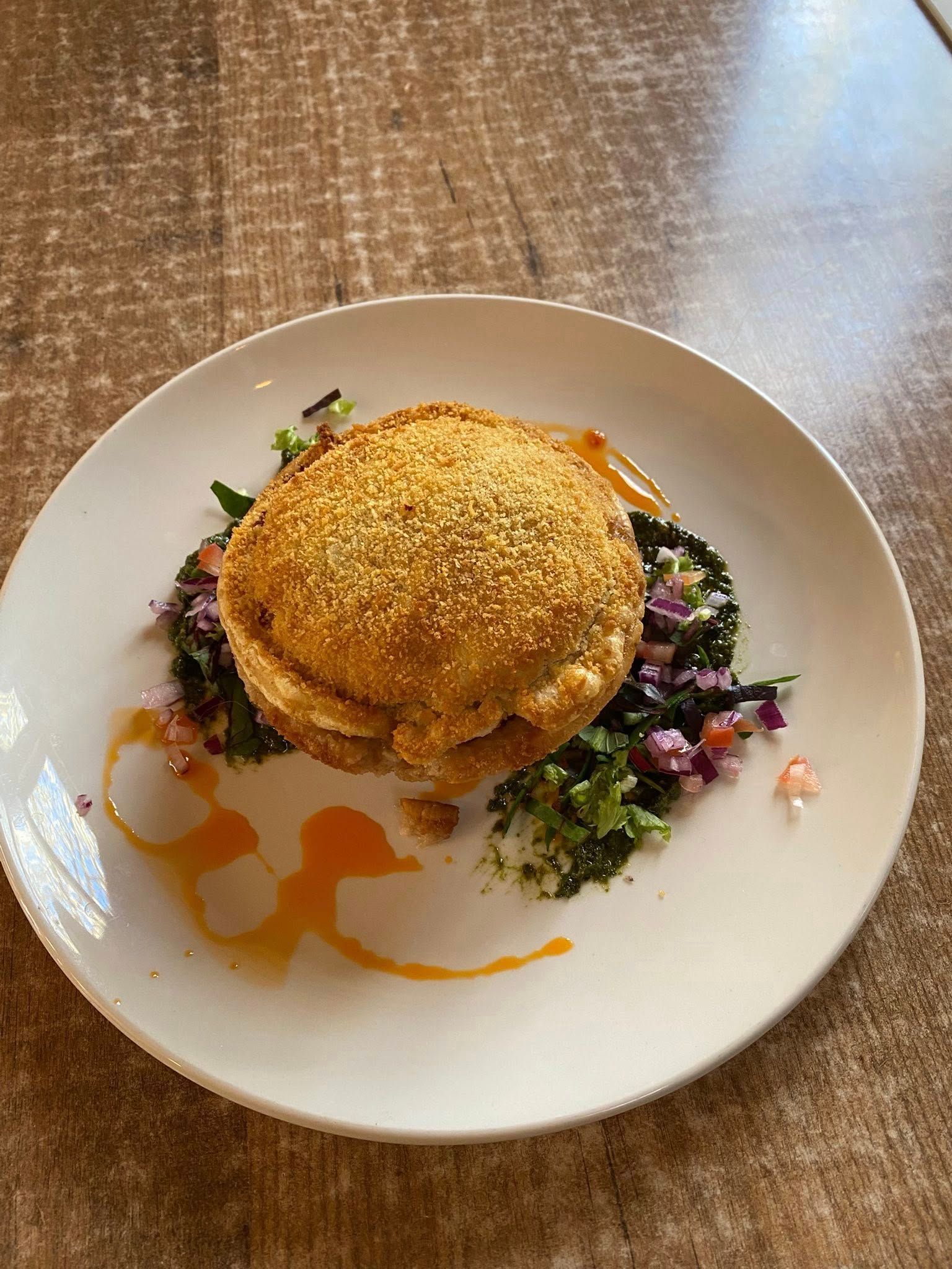 A golden-brown breaded patty sits on a bed of mixed greens with red accents, drizzled with orange sauce on a white plate. — 321 Cafe Learmonth In Learmonth, VIC