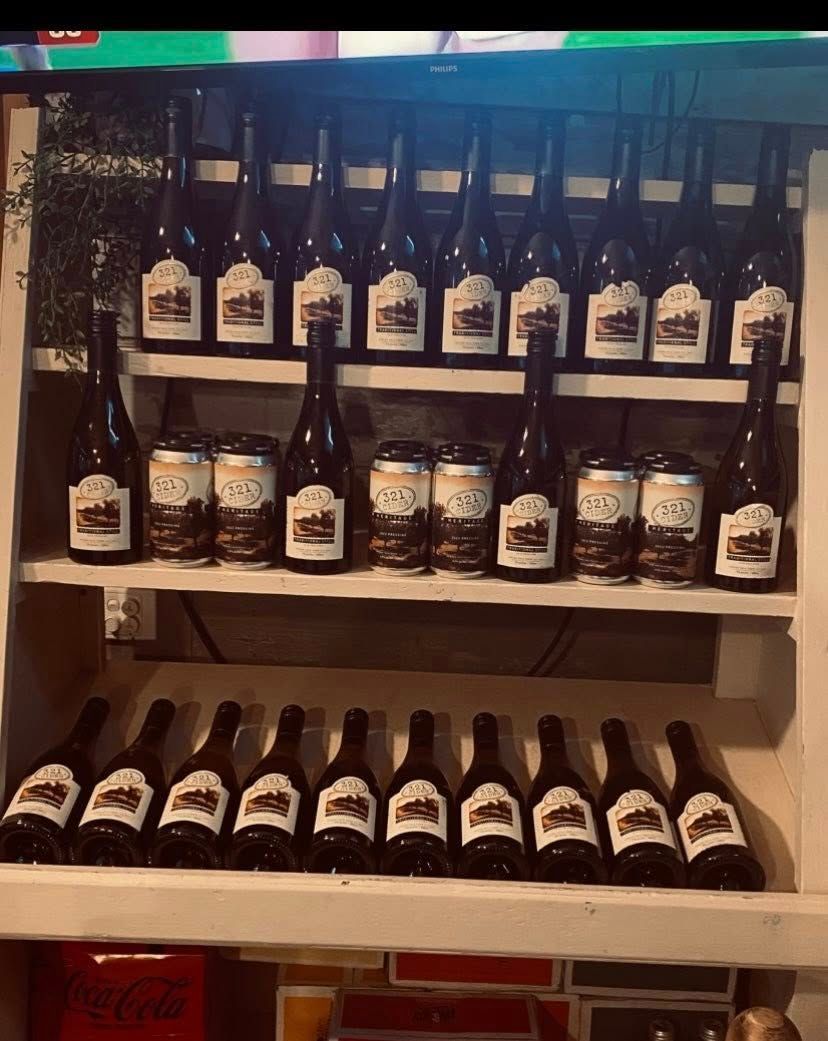 Three shelves hold rows of wine bottles and small drink cans in a dimly lit display. — 321 Cafe Learmonth In Learmonth, VIC