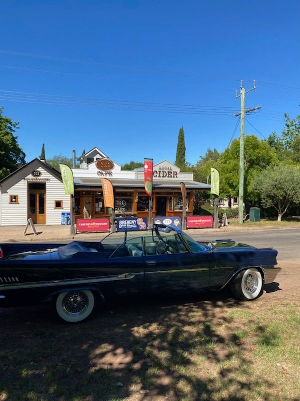 A dark-colored vintage convertible parked in front of a roadside cafe and cider house on a sunny day. — 321 Cafe Learmonth In Learmonth, VIC