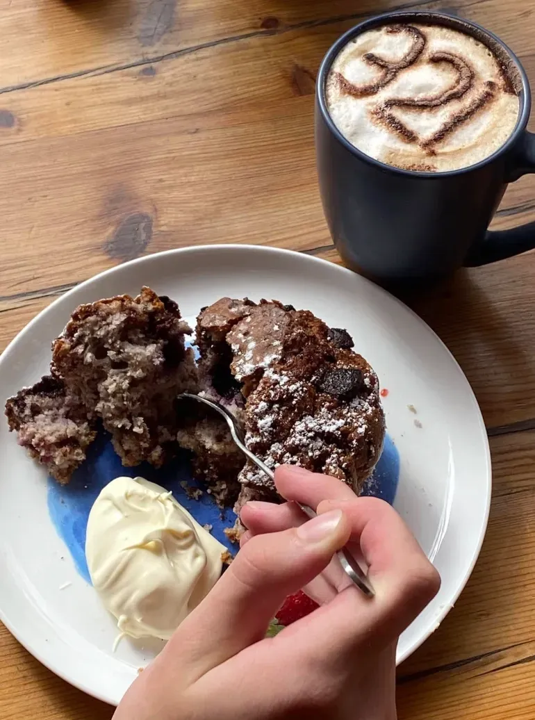A person uses a fork to eat a chocolate pastry served with cream on a white plate, next to a latte with 