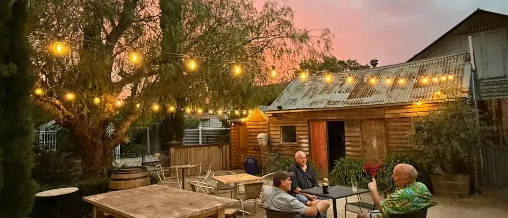 People Sitting at Tables Outside a Rustic Wooden Structure Under String Lights During a Sunset — 321 Cafe Learmonth In Learmonth, VIC