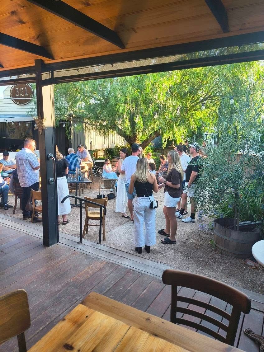 People socializing outdoors on a patio with wooden flooring, tables, and trees in the background. — 321 Cafe Learmonth In Learmonth, VIC