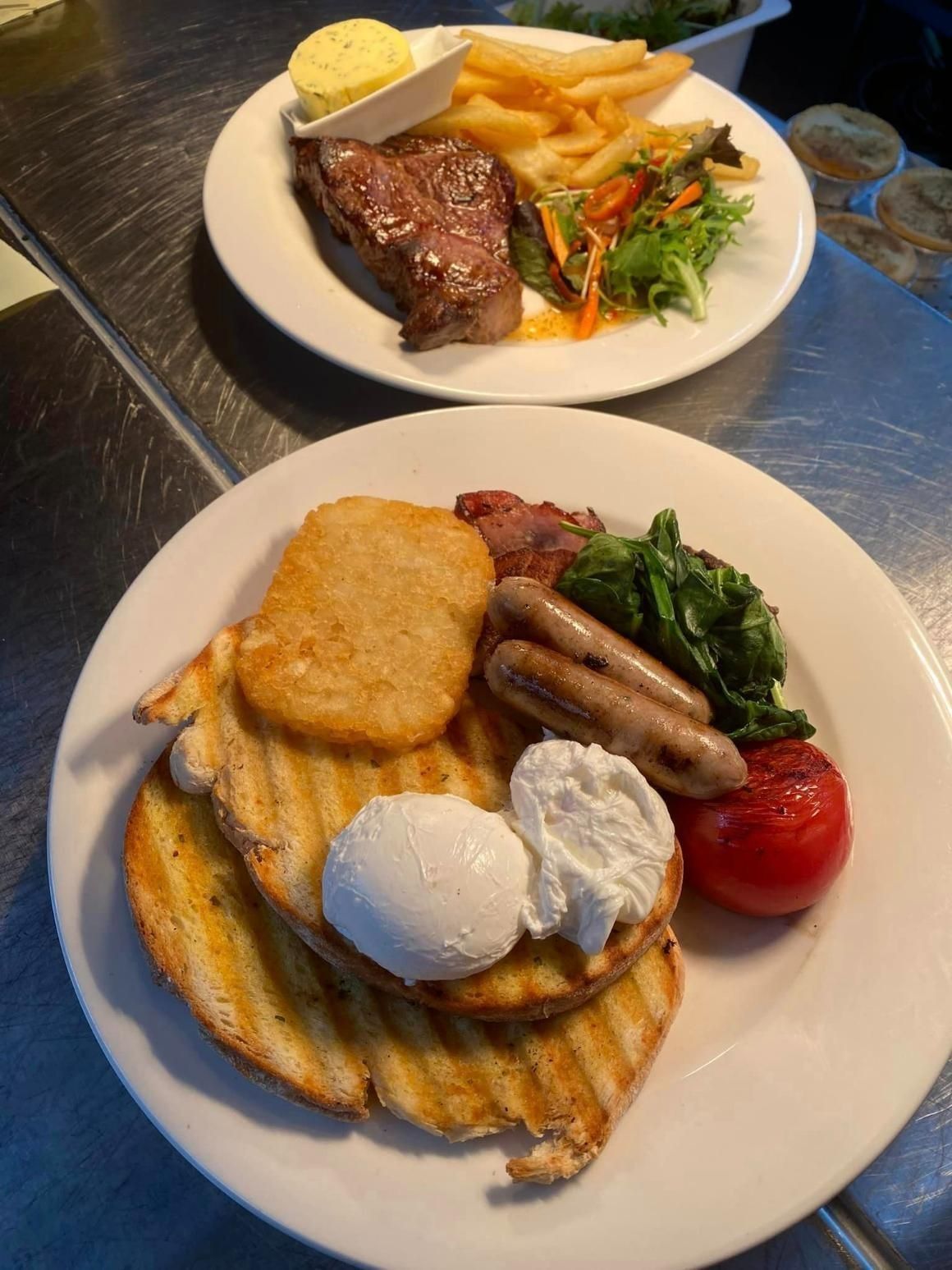Two plates of food on a metal counter, featuring a steak with fries and a full breakfast platter with eggs and sausage. — 321 Cafe Learmonth In Learmonth, VIC
