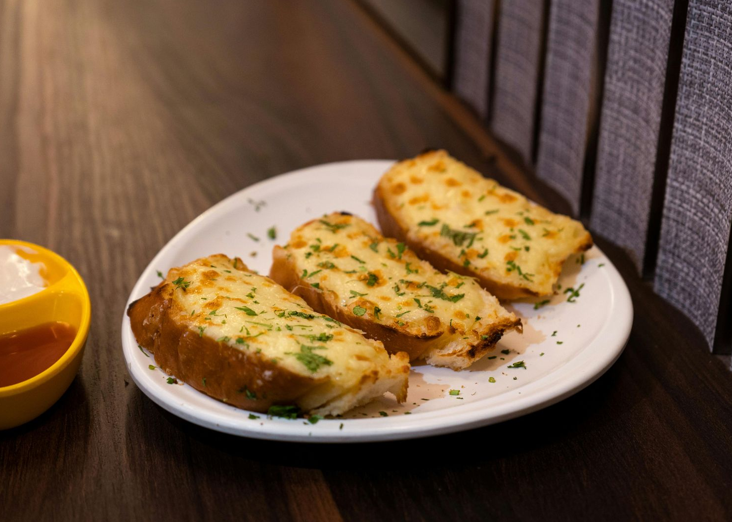 Three slices of toasted cheesy garlic bread on a white plate next to a small bowl with two dipping sauces on a table. — 321 Cafe Learmonth In Learmonth, VIC