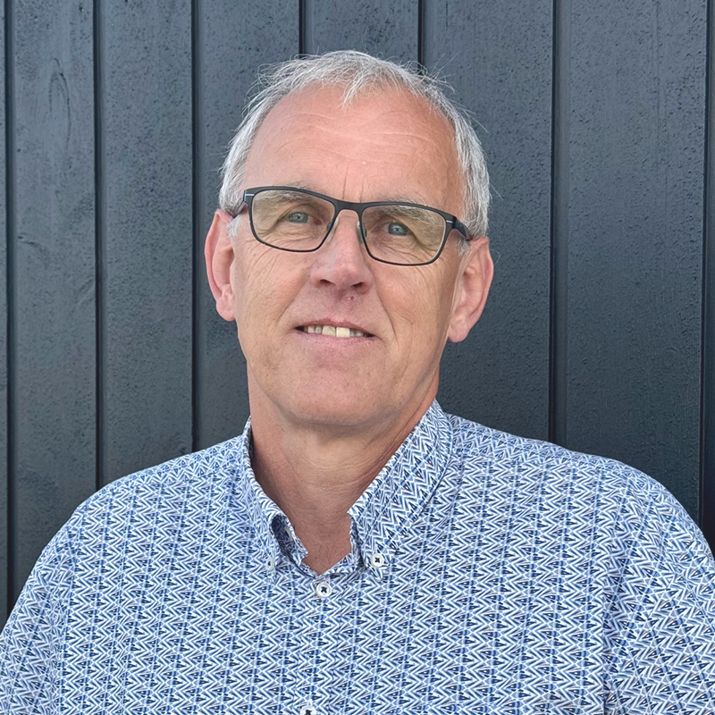 Mand med briller smiler, iført en mønstret blå skjorte, stående foran en blå/grøn væg.